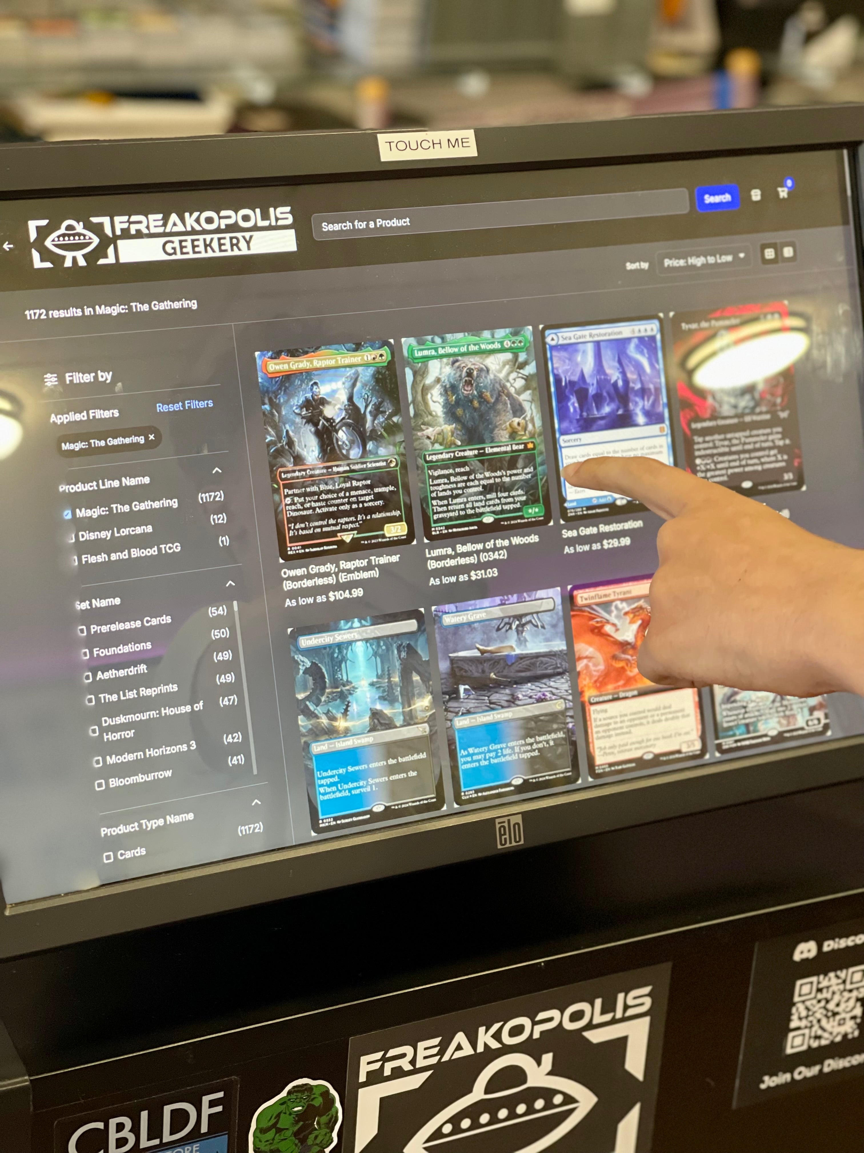 A hand makes trading card selections on a touch screen at the Greekery